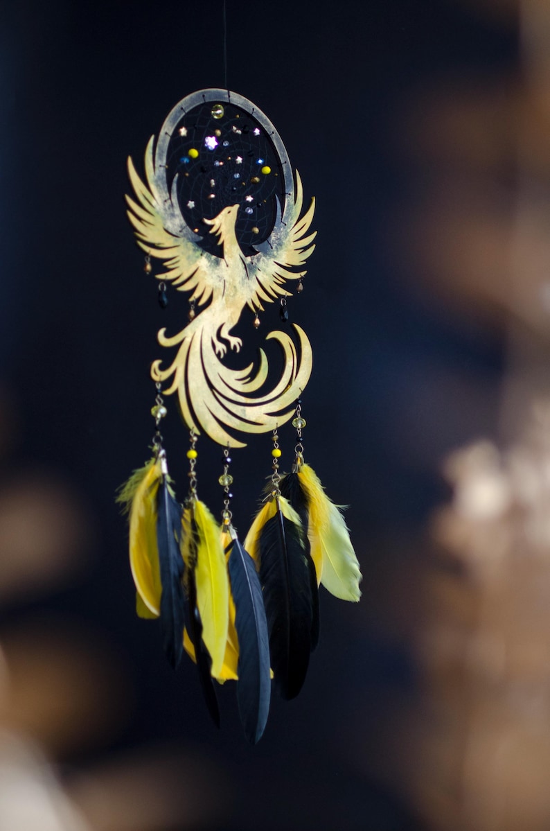 May include: A golden phoenix dreamcatcher with a black background. The phoenix is intricately cut with outstretched wings. Below, yellow and black feathers hang from beaded strands, adding a touch of color and movement to the design.
