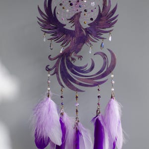 May include: A purple dreamcatcher featuring a phoenix design. The circular frame holds a web of string and beads. Below, purple and white feathers hang from the bottom. Clear crystal beads and purple beads are also included.