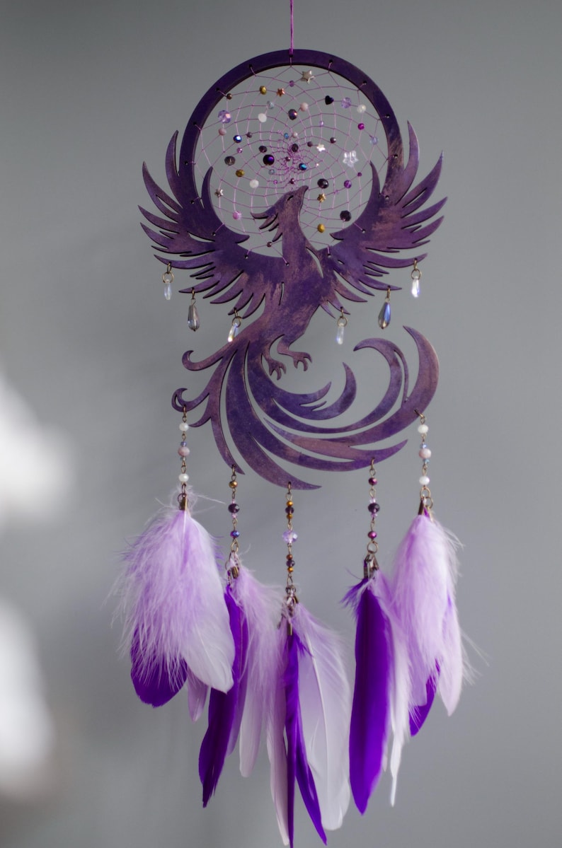 May include: A purple dreamcatcher featuring a phoenix design. The circular frame holds a web of string and beads. Below, purple and white feathers hang from the bottom. Clear crystal beads and purple beads are also included.