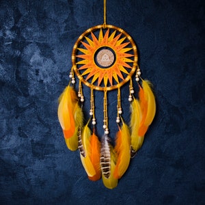 Op de afbeelding: Een gele en oranje droomvanger met witte kralen en veren. De droomvanger heeft een rond frame met een geweven ontwerp en een klein symbool in het midden.