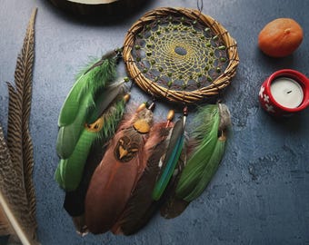 May include: A handmade dreamcatcher with a woven circular frame, adorned with green and brown feathers. A small candle in a red holder and a tangerine are also present. The dreamcatcher is a decorative item.
