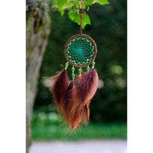 Puede incluir: Un atrapasueños verde y marrón con plumas marrones que cuelgan de él. El atrapasueños está colgado de una rama.