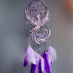 May include: A purple dreamcatcher featuring a phoenix design. The dreamcatcher has a circular frame with a web of string and decorative beads. Below the phoenix are hanging feathers in shades of purple and white.