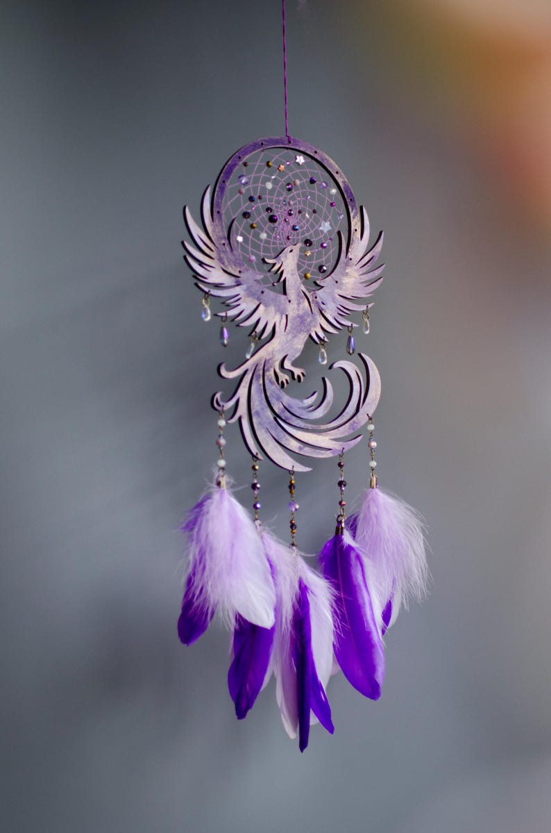 May include: A purple dreamcatcher featuring a phoenix design. The dreamcatcher has a circular frame with a web of string and decorative beads. Below the phoenix are hanging feathers in shades of purple and white.