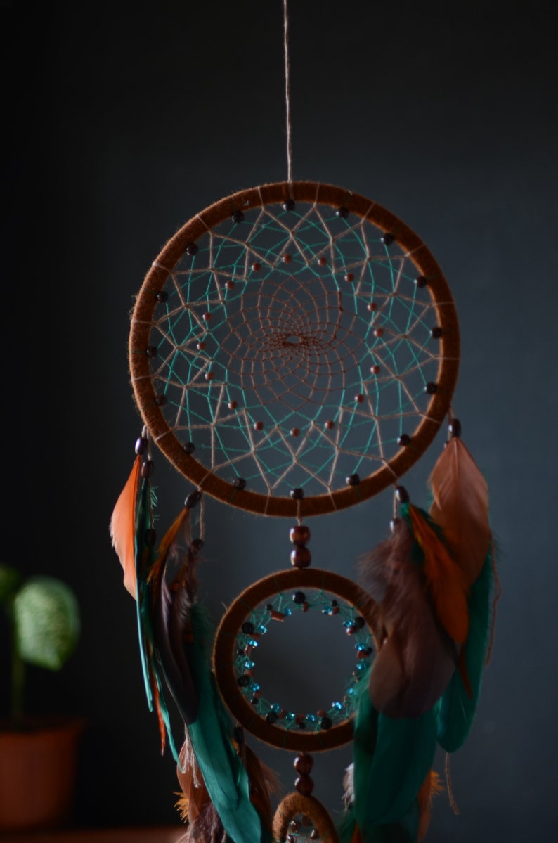 May include: A brown dreamcatcher with teal and brown feathers hanging from it. The dreamcatcher has two hoops, one larger than the other, and is decorated with beads.