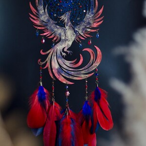 May include: A handcrafted dreamcatcher featuring a phoenix design. The circular frame and phoenix are painted in shades of red, blue, and white. Red and blue feathers hang below, with decorative beads and star accents. A celestial-themed wall hanging.