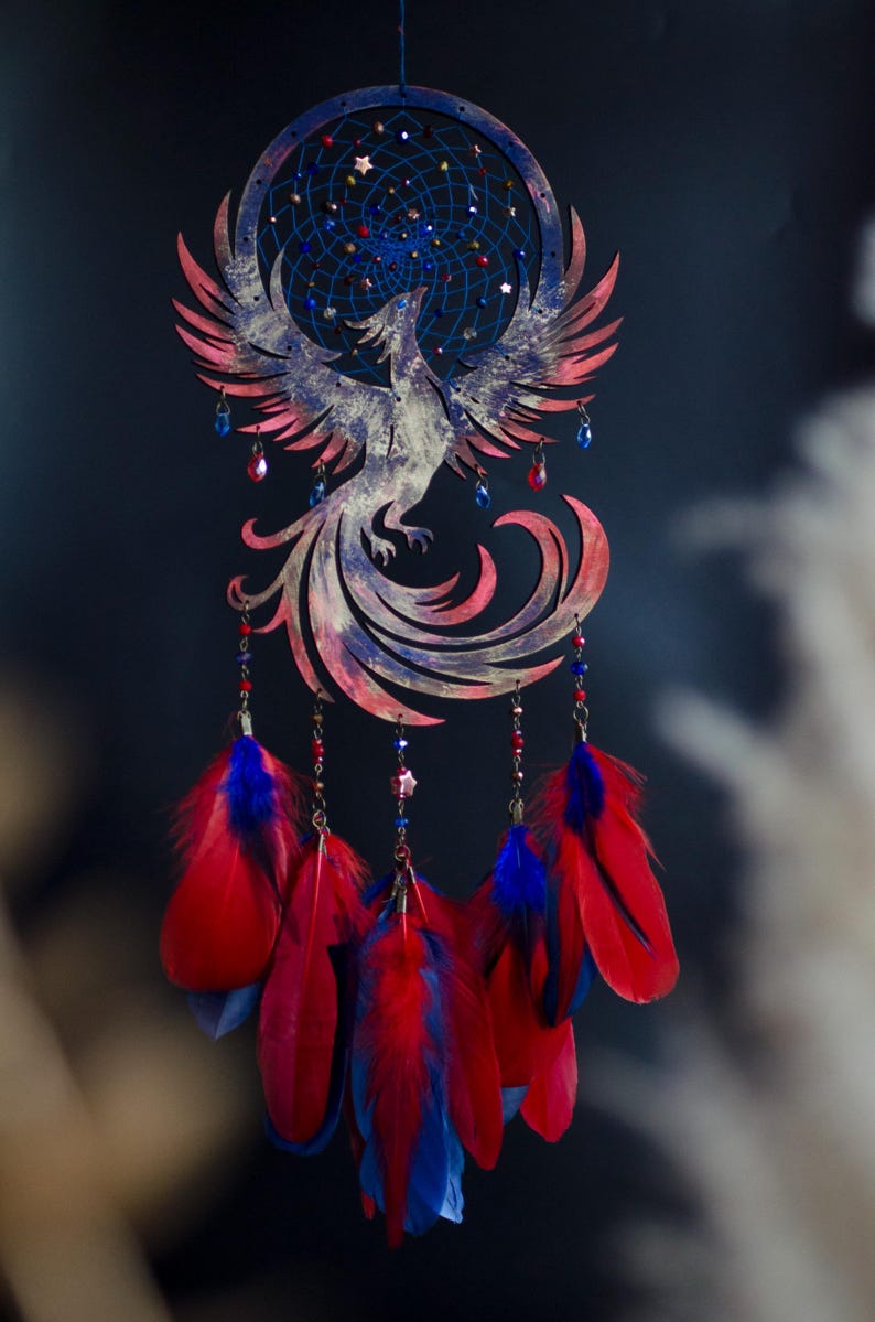 May include: A handcrafted dreamcatcher featuring a phoenix design. The circular frame and phoenix are painted in shades of red, blue, and white. Red and blue feathers hang below, with decorative beads and star accents. A celestial-themed wall hanging.