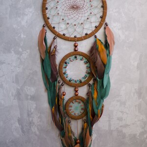 May include: A brown and green dreamcatcher with feathers and beads hanging from it. The dreamcatcher has three hoops, with the largest hoop at the top and the smallest hoop at the bottom.