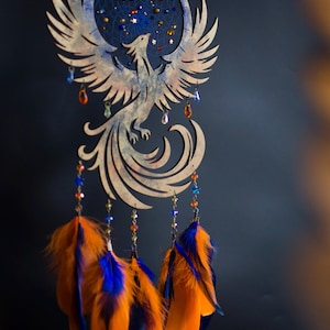 Handcrafted phoenix-shaped dreamcatcher with a deep blue woven web, orange and blue feathers, star beads, and crystal drops. Symbolizes rebirth and strength. Perfect for fantasy-themed or boho-style home decor.