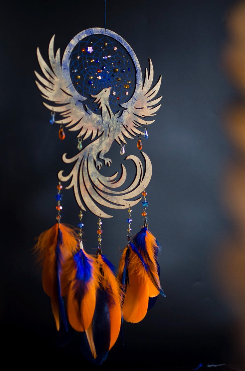 Handcrafted phoenix-shaped dreamcatcher with a deep blue woven web, orange and blue feathers, star beads, and crystal drops. Symbolizes rebirth and strength. Perfect for fantasy-themed or boho-style home decor.