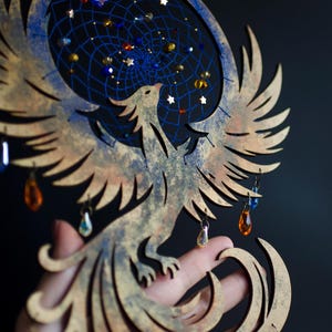 Handcrafted phoenix-shaped dreamcatcher with a deep blue woven web, orange and blue feathers, star beads, and crystal drops. Symbolizes rebirth and strength. Perfect for fantasy-themed or boho-style home decor.