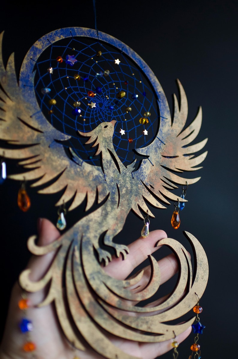 Handcrafted phoenix-shaped dreamcatcher with a deep blue woven web, orange and blue feathers, star beads, and crystal drops. Symbolizes rebirth and strength. Perfect for fantasy-themed or boho-style home decor.