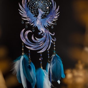 May include: A blue and white dreamcatcher featuring a phoenix design. The dreamcatcher has a circular web with beads and dangling feathers in shades of blue and white. The phoenix is intricately cut from wood.