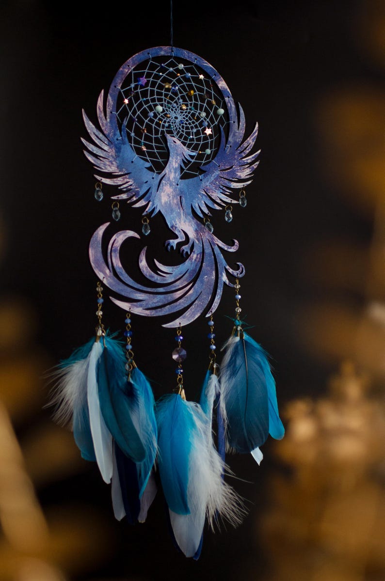 May include: A blue and white dreamcatcher featuring a phoenix design. The dreamcatcher has a circular web with beads and dangling feathers in shades of blue and white. The phoenix is intricately cut from wood.