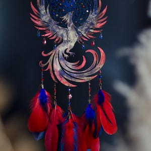 May include: A handcrafted dreamcatcher featuring a phoenix design. The circular frame and phoenix are painted in shades of red, blue, and white. Red and blue feathers hang below, with decorative beads and star accents. A celestial-themed wall hanging.