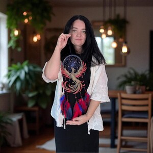May include: A handcrafted dreamcatcher featuring a phoenix design. The dreamcatcher has a dark blue web, red and gold accents, and red and blue feathers. The design is displayed against a black background.