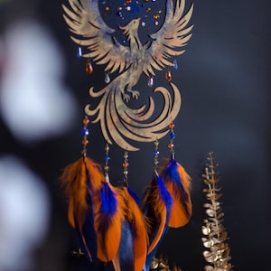 Handcrafted phoenix-shaped dreamcatcher with a deep blue woven web, orange and blue feathers, star beads, and crystal drops. Symbolizes rebirth and strength. Perfect for fantasy-themed or boho-style home decor.