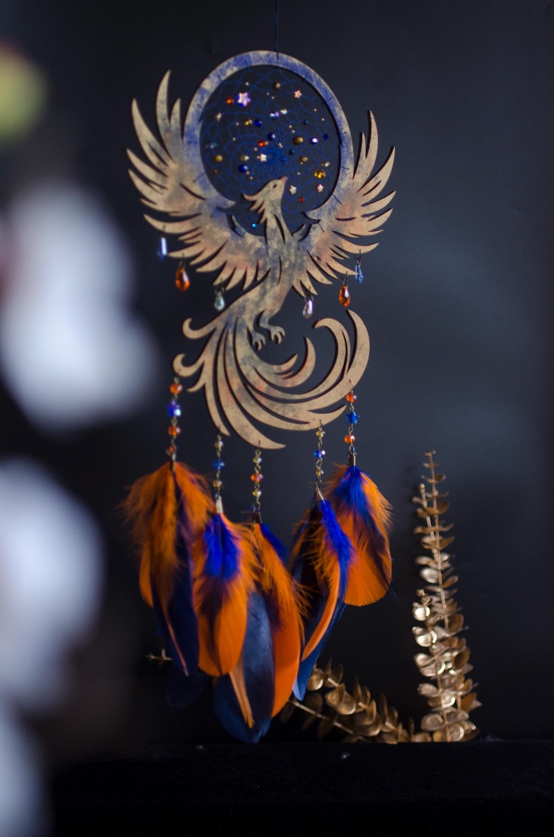 Handcrafted phoenix-shaped dreamcatcher with a deep blue woven web, orange and blue feathers, star beads, and crystal drops. Symbolizes rebirth and strength. Perfect for fantasy-themed or boho-style home decor.