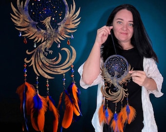 May include: A wooden dreamcatcher featuring a phoenix design with outstretched wings. The dreamcatcher has a blue and gold color scheme, with orange and blue feathers hanging below. Decorative beads and crystals are also included.