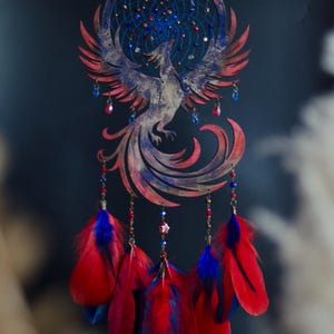 May include: A handcrafted dreamcatcher featuring a phoenix design. The circular frame and phoenix are painted in shades of red and blue. Strands of beads and feathers in red and blue hang below, creating a decorative wall hanging.