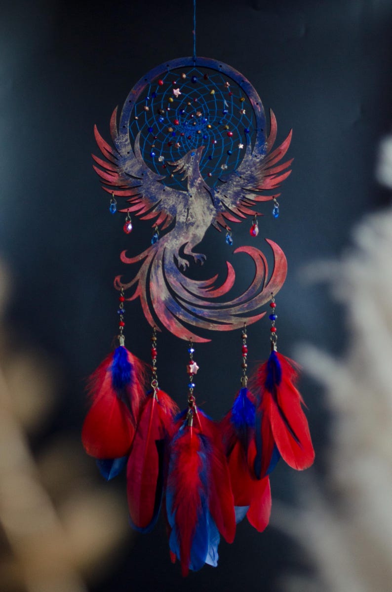 May include: A handcrafted dreamcatcher featuring a phoenix design. The circular frame and phoenix are painted in shades of red and blue. Strands of beads and feathers in red and blue hang below, creating a decorative wall hanging.