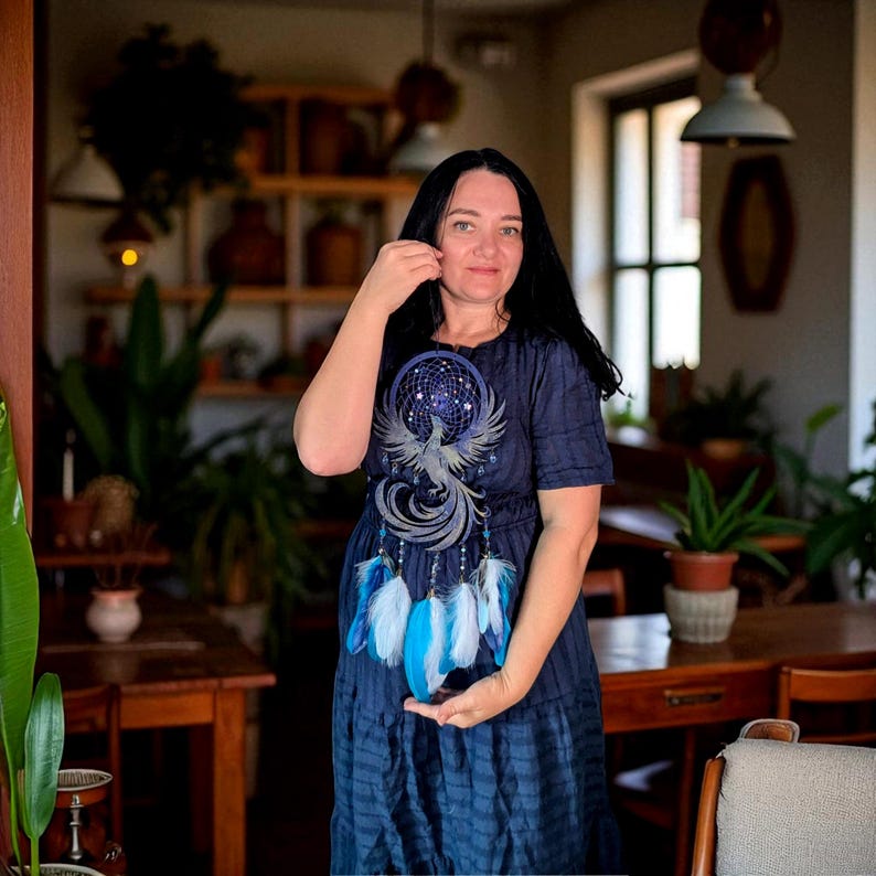 May include: A navy blue dress featuring a dreamcatcher design with a phoenix and blue and white feathers. The dress has short sleeves and a tiered skirt. The woman is holding the bottom of the dreamcatcher.