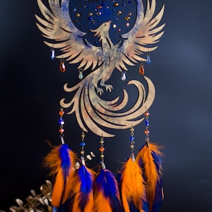 Handcrafted phoenix-shaped dreamcatcher with a deep blue woven web, orange and blue feathers, star beads, and crystal drops. Symbolizes rebirth and strength. Perfect for fantasy-themed or boho-style home decor.