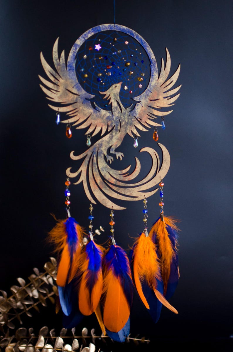 Handcrafted phoenix-shaped dreamcatcher with a deep blue woven web, orange and blue feathers, star beads, and crystal drops. Symbolizes rebirth and strength. Perfect for fantasy-themed or boho-style home decor.