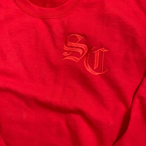 以下が含まれることがあります： 赤いスウェットシャツに、ゴシック体で「SC」が赤で刺繍されています