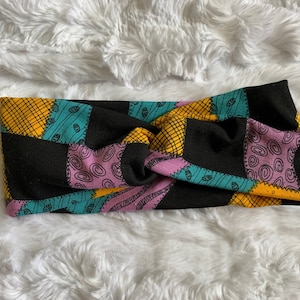 May include: A black, yellow, teal, and pink patchwork headband. The headband features a repeating pattern of squares with various designs, including swirls and stitched lines. The background is a soft, white, fluffy material.