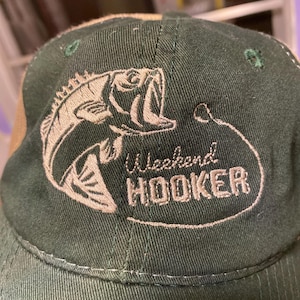 Può includere: Cappellino da baseball verde con un'immagine di pesce marrone e il testo "Weekend Hooker" ricamato sul davanti.