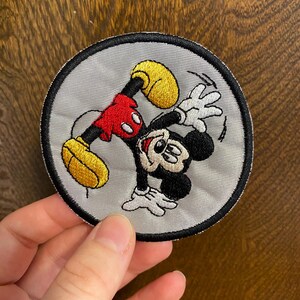 May include: Embroidered patch featuring Mickey Mouse in a dynamic pose. The patch is circular, with a gray background and a black border. Mickey is depicted in his classic colors: black, red, yellow, and white. The patch is likely for clothing or accessories.