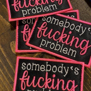 May include: Three black fabric patches with pink stitching. Each patch has the text "somebody's fucking problem" in white and pink lettering.
