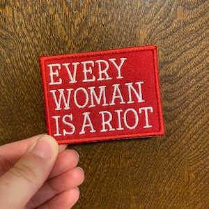 May include: Red fabric patch with white embroidered text that reads "EVERY WOMAN IS A RIOT".