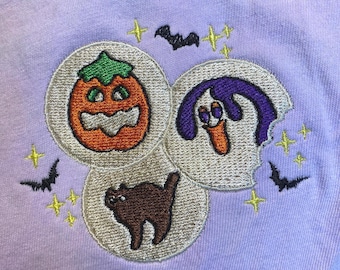 May include: A light purple fabric with embroidered Halloween-themed designs. The design includes a jack-o'-lantern, a ghost, and a black cat, each within a circular frame. Bats and stars are also embroidered.