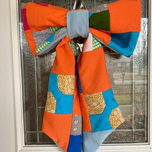 May include: A large, orange and blue patchwork bow with a variety of patterns and textures. The bow is tied with a light blue ribbon and is hanging on a white door.