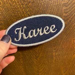 May include: An oval-shaped navy blue patch with the name "Karee" embroidered in white script. The patch has a white border and is likely designed to be sewn onto clothing or accessories.