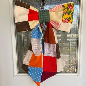 May include: A patchwork fabric bow with a variety of colors and patterns, including red, white, blue, green, orange, and brown. The bow is tied in a knot and hangs from a hook.