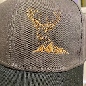 May include: A gray baseball cap with a black brim. The cap features an embroidered design of a deer with antlers above a mountain range in shades of brown. The cap has a structured fit.