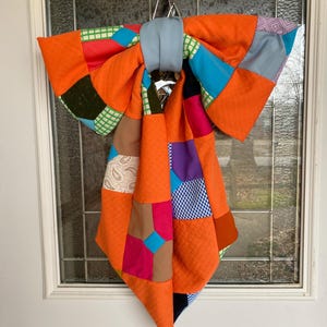 May include: A large orange bow made of patchwork fabric with various colors and patterns, hanging on a white door.