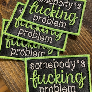May include: Four black fabric patches with green stitching and white text. The text on the patches reads "somebody's fucking problem".