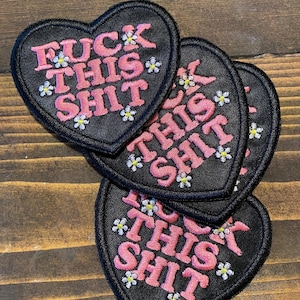 May include: A stack of black heart-shaped patches embroidered with the phrase "FUCK THIS SHIT" in pink, with small white flower accents. The patches are made of a textured fabric.