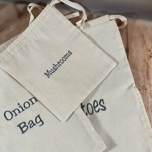 May include: Three cream-coloured canvas produce bags with black text. The bags are labelled "Mushrooms", "Onion Bag", and "Potatoes". Each bag has a loop handle. The bags are on a wooden surface.