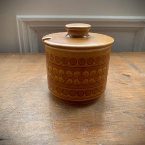 May include: A brown ceramic jar with a lid and a small handle. The jar has a cylindrical shape with a decorative pattern of circles and flowers around the body. The lid has a small knob and a slot for a spoon. The jar is sitting on a wooden surface.
