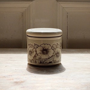 May include: A white ceramic jar with a floral design. The jar has a brown floral pattern with three flowers on the front. The jar is sitting on a light wood surface.