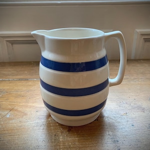 May include: A cream-coloured ceramic jug with blue horizontal stripes. The jug has a handle and a pouring spout. It is displayed on a wooden surface.