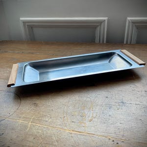 May include: A rectangular metal serving tray with a brushed silver finish. The tray has wooden handles on each end. The tray is sitting on a wooden surface.