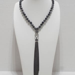 May include: A long black beaded necklace with a silver tassel pendant. The beads are round and have a dark grey colour with a slight shimmer. The tassel is made of small silver chains.