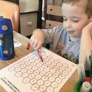 May include: A child uses a red marker to connect the numbers on a honeycomb-shaped game board. The board has a bee illustration at the bottom and the text "CAN YOU HELP BEE FIND HIS FRIEND BY FOLLOWING NUMBERS FROM 1-257".
