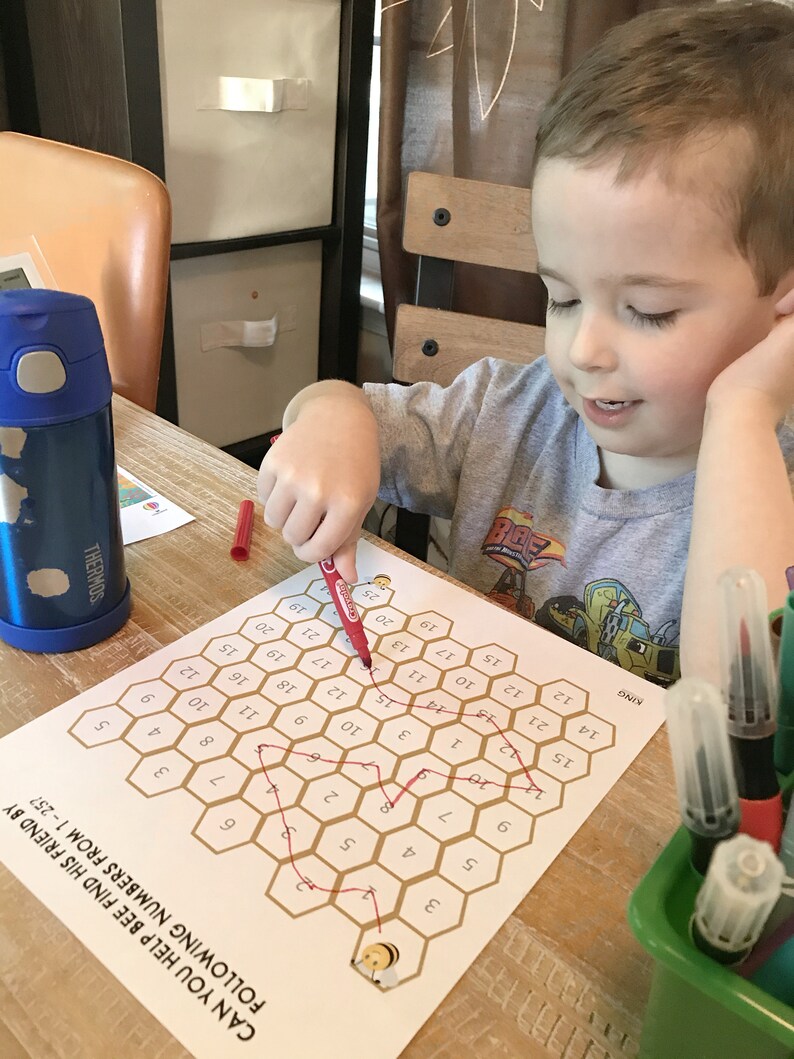 May include: A child uses a red marker to connect the numbers on a honeycomb-shaped game board. The board has a bee illustration at the bottom and the text "CAN YOU HELP BEE FIND HIS FRIEND BY FOLLOWING NUMBERS FROM 1-257".
